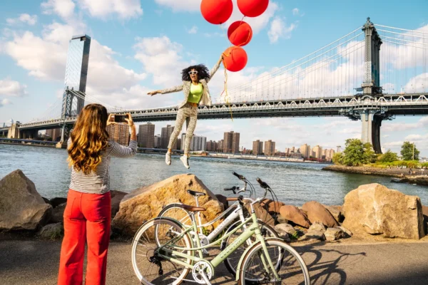 woman taking a photo of a woman flying up in balloons next to bicycles with a big bridge in the background